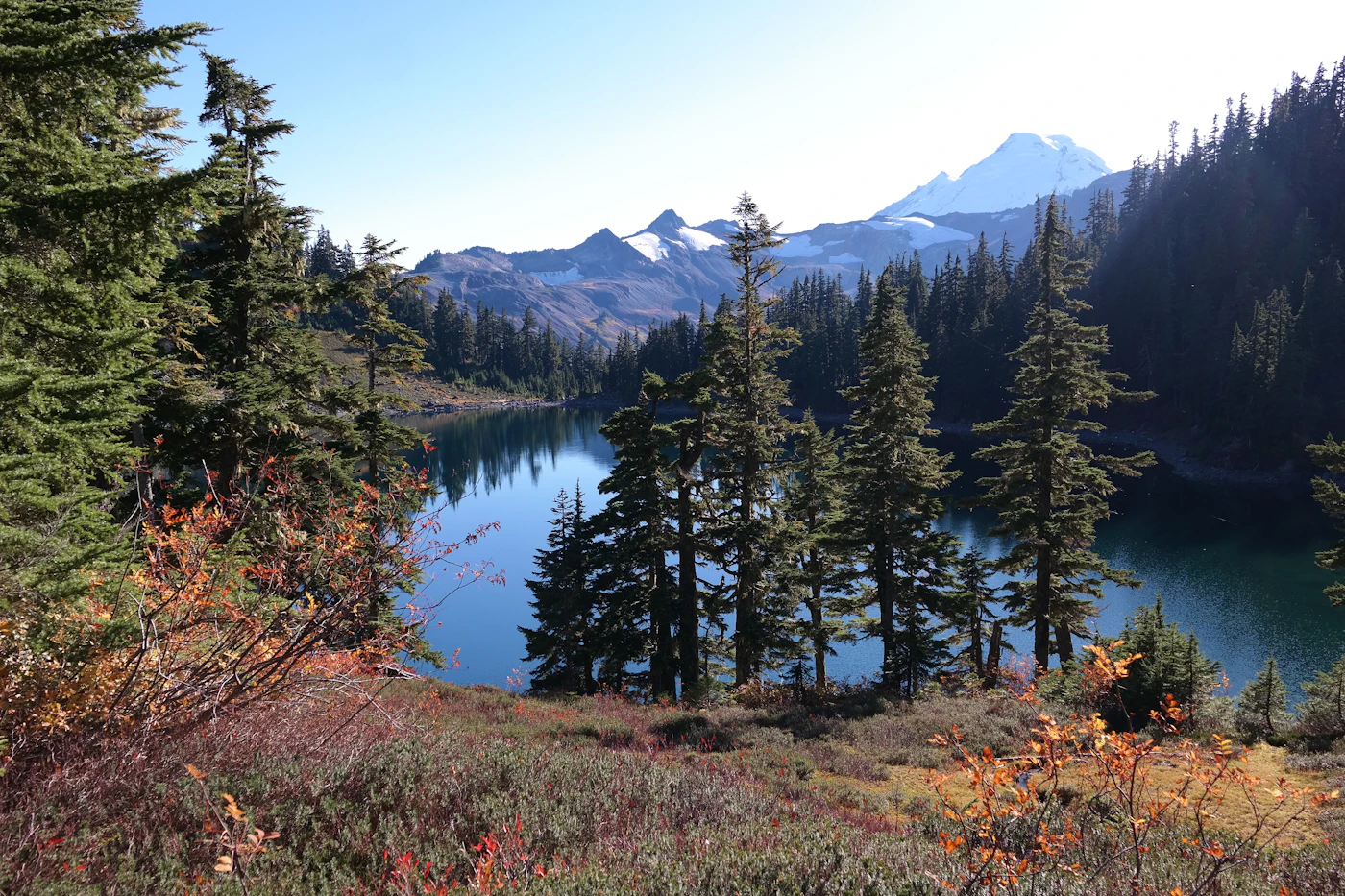 A still mountain lake reflects evergreen trees, with snow on the slopes beyond.